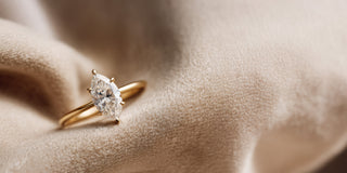A Réia yellow gold solitaire ring with a marquise-cut lab-grown diamond in a six-prong setting, resting on beige fabric.