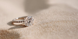 A Réia white gold engagement ring featuring a round lab-grown diamond on a pavé band, resting on sand.