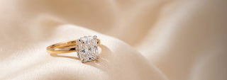 A Réia yellow gold solitaire engagement ring with a Cushion-cut lab-grown diamond in a four-prong setting, displayed on flowing satin fabric.
