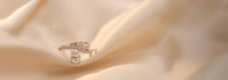 A Réia rose gold ring with an oval lab-grown diamond center stone and marquise and round diamond floral accents, resting on beige fabric.