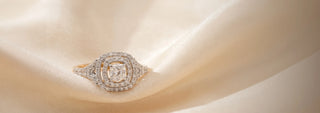 A Réia white gold engagement ring featuring a square center diamond with a double diamond halo and pavé band, resting on beige fabric.