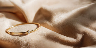 Réia yellow gold simple bangle with a single bezel-set lab-grown diamond, resting on beige fabric.