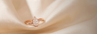 A Réia yellow gold engagement ring with a marquise-cut lab-grown diamond center stone and diamond accents, resting on beige fabric with dried flowers.