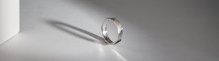 A Réia white gold band ring with a modern, angular, and faceted design, is displayed on a gray surface with strong shadows.