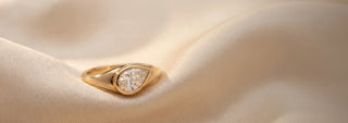 Réia yellow gold engagement ring featuring a pear-cut lab-grown diamond on a pavé band, set on champagne fabric.