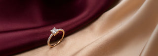 A Reia yellow gold engagement ring with a princess cut lab-grown diamond is displayed on folded beige velvet near small dried flowers.