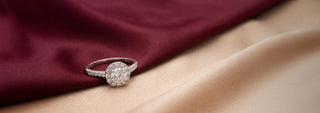 A Réia white gold engagement ring featuring a Cushion-cut lab-grown diamond with a diamond halo and pavé band, resting on beige fabric.