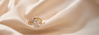 A Réia yellow gold engagement ring with an emerald cut lab-grown diamond and floral diamond side accents, resting on beige fabric with dried flowers.