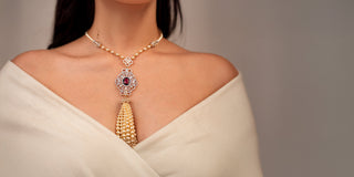 A Réia gold and pearl statement necklace featuring a large ornamental pendant with a dark red stone, white lab-grown diamonds, and a hanging tassel of gold beads, worn on a model's neck.