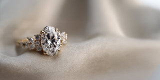  A Réia yellow gold cocktail engagement ring with an oval lab-grown diamond center stone and diamond side accents, resting on  beige fabric.