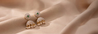 Front angled view of 14KT Yellow Gold diamond jhumka earrings featuring emerald center stones, floral diamond tops, and pearl drops. Displayed on a beige satin backdrop.