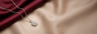Angled view of a 14KT White Gold floral diamond pendant necklace featuring a flower-shaped cluster of round diamonds on a delicate chain. Displayed on burgundy and beige satin.