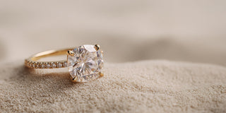 A Réia yellow gold solitaire engagement ring with a Cushion-cut lab-grown diamond and a pavé diamond band, resting on sand.