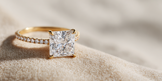 A Reia yellow gold engagement ring features a large square-cut lab-grown diamond on a pavé band, resting on fine pale beach sand.