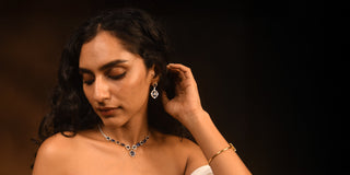 A model wearing a Réia white gold necklace and matching drop earrings set with blue stones and lab-grown diamonds, and a gold bangle, against a dark background.