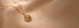 Angled view of a 14KT Yellow Gold floral pendant necklace featuring multicolored gemstones, diamond accents, and a scalloped halo. Displayed on a beige satin backdrop, highlighting its intricate design.