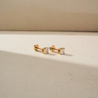 Side view of solitaire diamond stud earrings in yellow gold, featuring round brilliant-cut diamonds in secure prong settings. Displayed on a beige backdrop, the design highlights timeless simplicity.