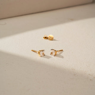 Angled view of solitaire diamond stud earrings in yellow gold, featuring round brilliant-cut diamonds in prong settings. Displayed on a beige backdrop, the minimalist design offers subtle sparkle.