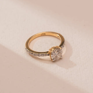 Top perspective view of Reia’s 14 KT Yellow Gold Eternal Glow Diamond Solitaire Ring. The round diamond and pavé band sit on a soft pink surface, showing off its warm, symmetrical shine.