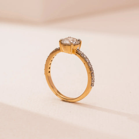 Side view of Reia’s 14 KT Yellow Gold Eternal Glow Diamond Solitaire Ring. The round diamond is held high in a simple gold setting on a soft pink surface, showing its bright and clean design.