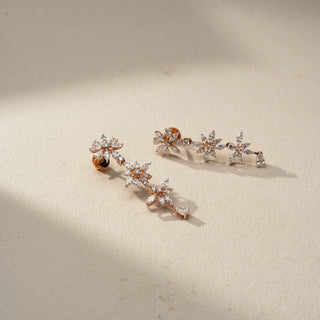 Angled view of floral diamond drop earrings in rose gold, featuring marquise-cut stones arranged in blooming flower motifs. Set against a beige backdrop, the elegant design offers delicate sparkle.