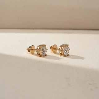 Side view of round brilliant diamond stud earrings in yellow gold basket settings with screw-back posts, displayed on a soft neutral surface with warm lighting and subtle shadows.