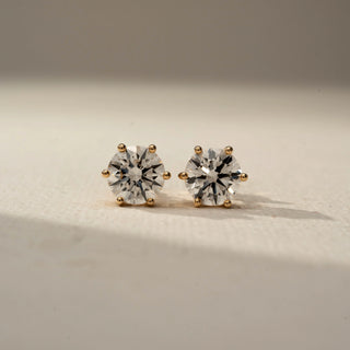 Front view of round brilliant diamond stud earrings in classic six-prong yellow gold settings, displayed on a soft neutral surface with warm lighting and subtle shadows.