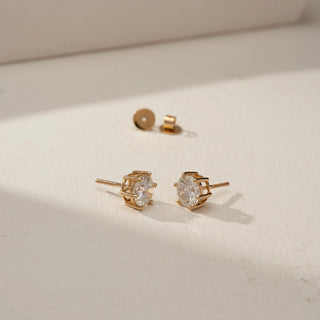 Angled side view of round brilliant diamond stud earrings in yellow gold basket settings, displayed on a neutral surface with screw-back closures and warm natural lighting.