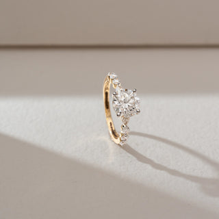 Side angled view of solitaire diamond ring in yellow gold, featuring a round lab-grown diamond at the centre with marquise and round diamond accents along the band. Set on a soft neutral backdrop, it highlights brilliance.