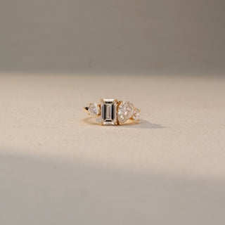 Front view of a three-stone engagement ring in 14 KT Yellow Gold, featuring an emerald-cut lab-grown diamond flanked by pear-shaped and round side diamonds, displayed against a beige backdrop.