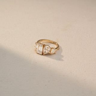 Side view of a three-stone engagement ring in 14 KT Yellow Gold, featuring an emerald-cut lab-grown diamond flanked by pear-shaped and round side diamonds, displayed against a beige backdrop.