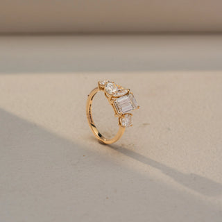 Angled view of a three-stone engagement ring in 14 KT Yellow Gold, featuring an emerald-cut lab-grown diamond flanked by pear-shaped and round side diamonds, displayed against a beige backdrop.