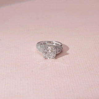 Close-up view of a cushion diamond ring in 14KT White Gold, featuring a pavé-set split band against a soft pink backdrop.