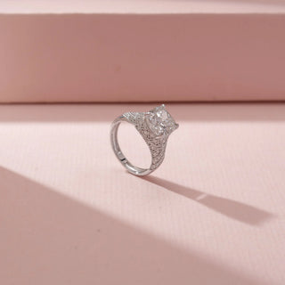 Angled view of a cushion  diamond ring in 14KT White Gold, featuring a pavé-set split band against a soft pink backdrop.