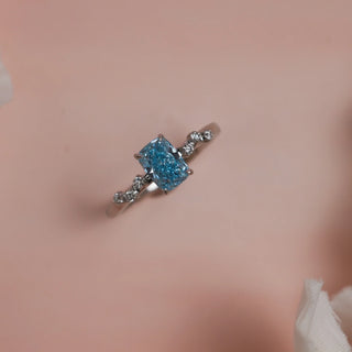 Top view of Reia Ocean Mist Cushion Diamond Ring on a soft blush-peach background, featuring a cushion-cut blue lab-grown diamond with six white accent stones on a 14KT white gold band.