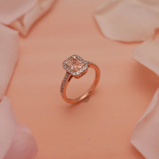 Angled top view of Reia Rose Halo Radiant Diamond Ring on a peach background with soft rose petals, highlighting the blush pink radiant-cut centre stone and halo of EF VVS diamonds.