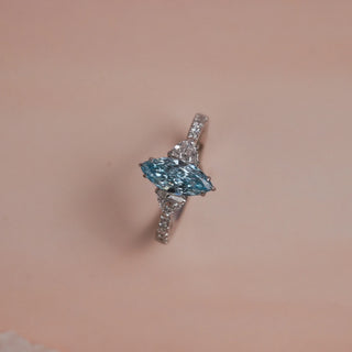 Top view of Reia Aqua Trine Marquise Diamond Ring on a blush-peach background, featuring a marquise-cut blue lab-grown diamond flanked by trilliant side stones and pave diamonds on a 14KT white gold band.