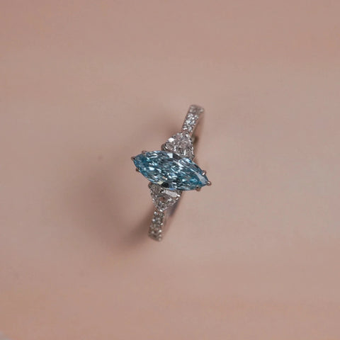 Top view of Reia Aqua Trine Marquise Diamond Ring on a blush-peach background, featuring a marquise-cut blue lab-grown diamond flanked by trilliant side stones and pave diamonds on a 14KT white gold band.