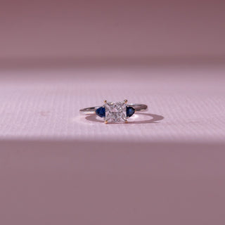 Front view of a diamond ring in white gold, featuring a princess-cut center diamond with blue pear-shaped side stones, set against a soft pink backdrop highlighting contrast and brilliance.
