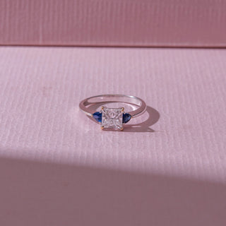 Top view of a diamond ring in white gold, featuring a princess-cut center diamond with blue pear-shaped side stones, set on a soft pink surface highlighting contrast and brilliance.
