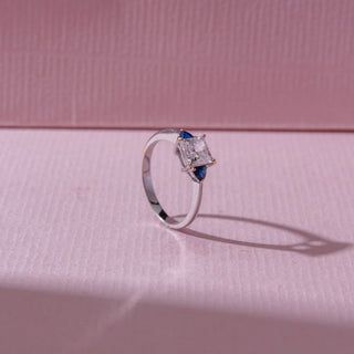 Angled view of a diamond ring in white gold, featuring a princess-cut center diamond with blue pear-shaped side stones, set on a soft pink surface highlighting depth and brilliance.