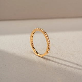 Angled view of a diamond eternity band in 14 KT Yellow Gold, featuring a continuous row of round lab-grown diamonds in a classic shared-prong setting, displayed against a beige backdrop.