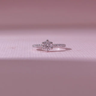 Front view of a white gold diamond ring, featuring a round solitaire center stone with pavé-set band detailing. Captured against a soft pink backdrop, the design reflects timeless sparkle and elegance.