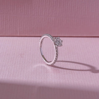 Side view of a white gold diamond ring, featuring a round solitaire center stone with pavé-set band detailing. Captured against a soft pink backdrop, the design reflects timeless sparkle and elegance.