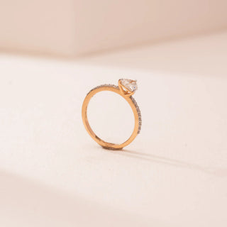 Side view of Reia’s 18 KT Yellow Gold Forever Heart Ring. The heart-shaped diamond is held in an elevated setting on a pavé-set gold band, shown against a soft pink surface.