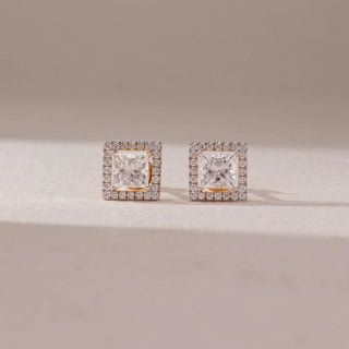 Front view of diamond stud earrings in 14KT yellow gold, featuring princess-cut center diamonds surrounded by square pavé halo settings, displayed on a beige surface highlighting brilliance and symmetry.
