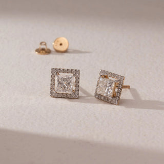 Angled view of diamond stud earrings in 14KT yellow gold, featuring princess-cut center diamonds with square pavé halo settings and screw backs, set on a beige surface highlighting brilliance and detail.
