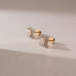 Side view of diamond stud earrings in 14KT yellow gold, featuring princess-cut center diamonds with square pavé halo settings and screw backs, displayed on a beige surface highlighting depth and sparkle.
