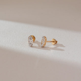 Angled view of marquise diamond stud earrings in white gold, featuring elongated lab-grown diamonds in a prong setting. Set against a soft beige textured background with gentle shadows.