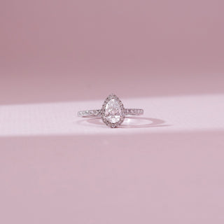 Front view of the Pear Halo Radiant Diamond Ring in 14KT white gold, featuring a pear-shaped center diamond encircled by a sparkling halo with a delicate pavé diamond band on a soft pink background.